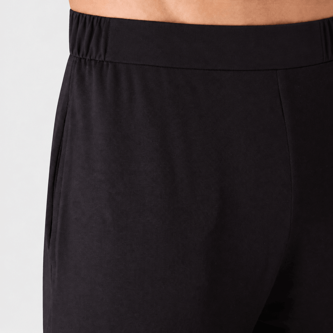 Close-up of a person wearing black ounge pants, showing the elastic waistband and side seam against a light gray background.